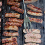 Close-up of sausage links grilling on a barbecue grate with smoky steam and a metal turning fork nearby.