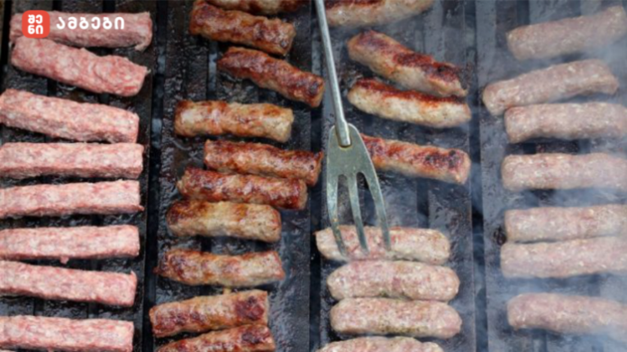 Close-up of sausage links grilling on a barbecue grate with smoky steam and a metal turning fork nearby.
