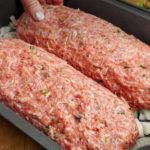 Two raw seasoned ground beef loaves in a pan over chopped onions, ready to bake, with herbs visible in the meat mixture