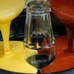 Colorful paint being poured into a round mold from left yellow and right orange-red streams meeting near a glass bottle in the center.