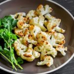 Plate of roasted cauliflower florets with a greens garnish on a dark wooden table, beside a decorative fork.
