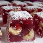 Red jelly dessert squares with banana pieces inside, coated in shredded coconut, arranged on a tray.