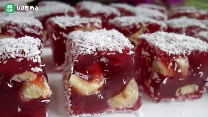Red jelly dessert squares with banana pieces inside, coated in shredded coconut, arranged on a tray.