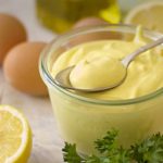 Glass jar of creamy lemon mayonnaise with a spoon, surrounded by lemons, eggs, and parsley on a kitchen counter.