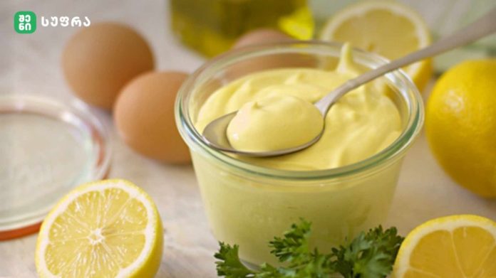 Glass jar of creamy lemon mayonnaise with a spoon, surrounded by lemons, eggs, and parsley on a kitchen counter.