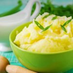 Green bowl of creamy mashed potatoes garnished with chives on a turquoise wooden table.