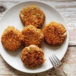 Five crispy breaded croquettes arranged on a white plate atop a rustic wooden table, with a fork nearby and a logo in the corner.