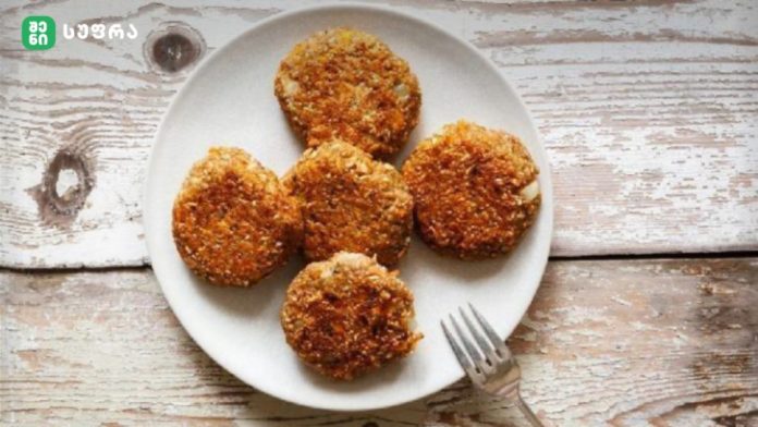 Five crispy breaded croquettes arranged on a white plate atop a rustic wooden table, with a fork nearby and a logo in the corner.
