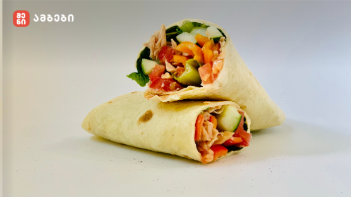 Two fresh wraps filled with cucumber, tomatoes, peppers, greens and pink shredded protein, stacked on a white surface; red logo watermark in the upper-left corner.