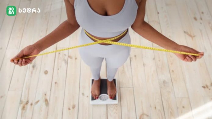 Top-down view of a person measuring their waist with a yellow tape while standing on a scale on a wooden floor.