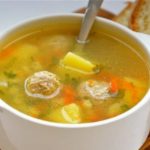 White ceramic bowl of vegetable soup with carrots, potatoes and herbs, served with slices of bread on a wooden board.