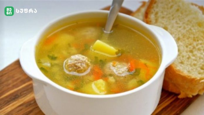 White ceramic bowl of vegetable soup with carrots, potatoes and herbs, served with slices of bread on a wooden board.