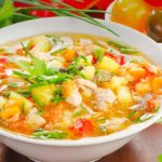 Bowl of hearty vegetable soup with potatoes, carrots, and herbs on a wooden table, garnished with parsley.