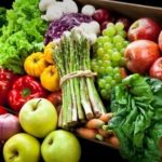 Assorted fresh fruits and vegetables arranged in a wooden crate, including bananas, apples, peppers, leafy greens, grapes, and bunches of asparagus.