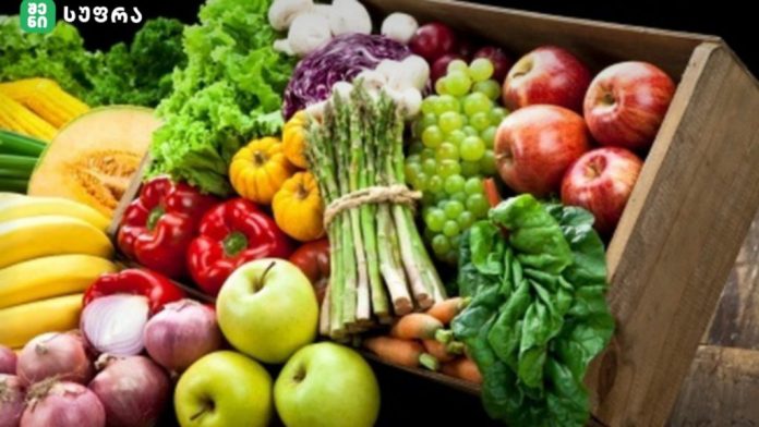 Assorted fresh fruits and vegetables arranged in a wooden crate, including bananas, apples, peppers, leafy greens, grapes, and bunches of asparagus.
