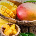 Mango cut into cubes in a wooden bowl beside a whole mango on a rustic wooden surface with green leaves nearby