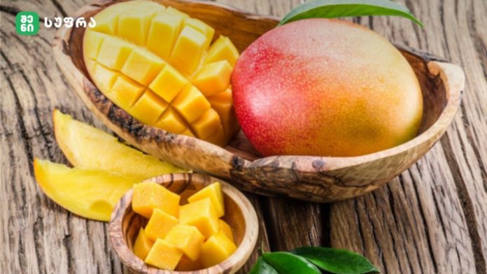 Mango cut into cubes in a wooden bowl beside a whole mango on a rustic wooden surface with green leaves nearby