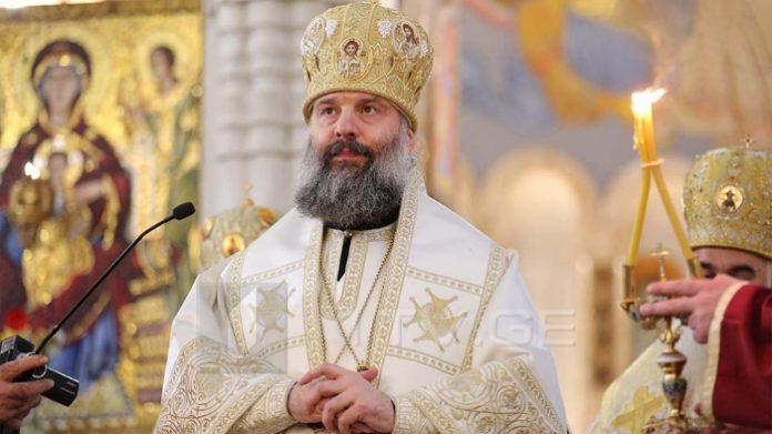 Orthodox bishop in gold mitre and white liturgical vestments, standing in a church with icons nearby.