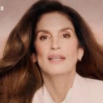 Portrait of a mature woman with long brown hair wearing a white blazer, posed against a peach background with a small logo in the top-left corner.