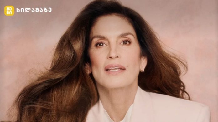 Portrait of a mature woman with long brown hair wearing a white blazer, posed against a peach background with a small logo in the top-left corner.