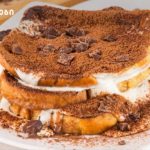 Layered tiramisu-style dessert with cream between bread slices, cocoa powder and chocolate chunks on top