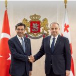 Two men in suits shake hands in a formal setting, with Georgian and Turkish flags and a crest behind them.