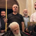 Five men stand behind an elderly man with a white beard lying in a coffin inside a church setting; one man holds a long yellow staff.