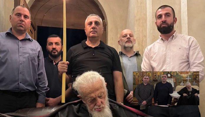 Five men stand behind an elderly man with a white beard lying in a coffin inside a church setting; one man holds a long yellow staff.