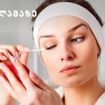 Woman with a white headband applies makeup to her eye using a cotton swab, in a bathroom setting with a logo in the corner.