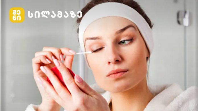 Woman with a white headband applies makeup to her eye using a cotton swab, in a bathroom setting with a logo in the corner.