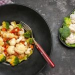 Black bowl filled with a vegetable medley (broccoli, cauliflower, carrots) and a red-handled fork; a separate plate shows broccoli and cauliflower florets.