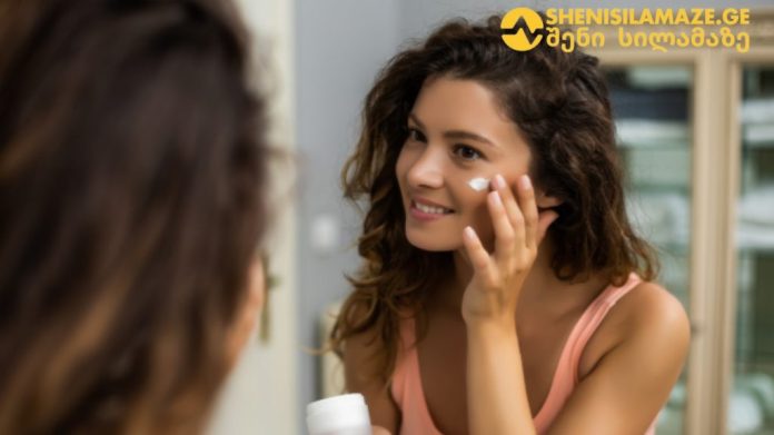 Woman applying facial cream to her cheek while looking in a mirror, skincare routine at home