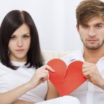 Two adults sit on a sofa, holding a torn red paper heart between them, looking unhappy.