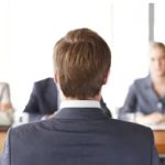 Job interview: a candidate in a suit seated with his back to the camera, facing three interviewers at a table.