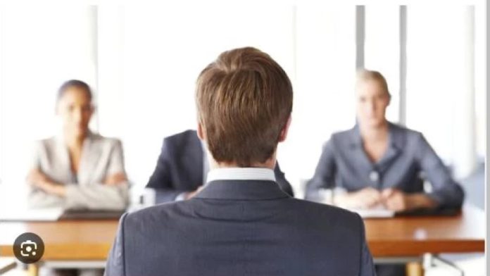 Job interview: a candidate in a suit seated with his back to the camera, facing three interviewers at a table.