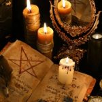 Open spellbook with a pentagram drawn inside, a feather quill, and several lit candles on a dark ceremonial altar with a decorative mirror and crystals nearby.
