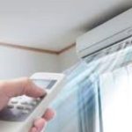 Person holds a remote control aimed at a wall-mounted air conditioner unit above a window with sheer curtains in a home setting.