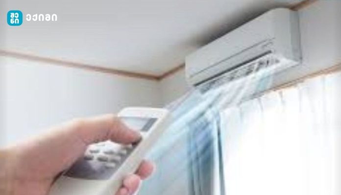Person holds a remote control aimed at a wall-mounted air conditioner unit above a window with sheer curtains in a home setting.