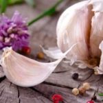 Garlic bulb with a peeled clove on a weathered wooden surface, with a purple flower in the background.