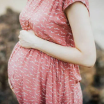 Pregnant woman in a pink patterned dress gently cradling her baby bump outdoors.
