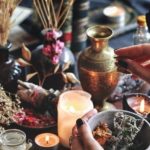 Hands arranging dried herbs on a candlelit altar with vases and bowls nearby, creating a ritual scene.