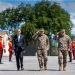 Civilian official in a dark suit walking between two soldiers in camouflage as red-uniform ceremonial guards stand at attention outdoors during a military honor ceremony.