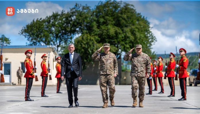 Civilian official in a dark suit walking between two soldiers in camouflage as red-uniform ceremonial guards stand at attention outdoors during a military honor ceremony.