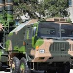 Camouflage military missile launcher on a large six‑wheel truck displayed outdoors in a yard.
