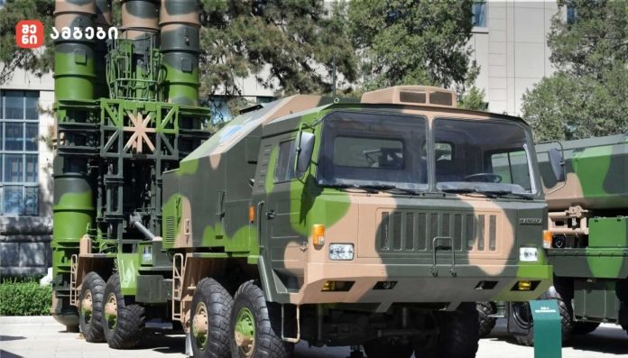 Camouflage military missile launcher on a large six‑wheel truck displayed outdoors in a yard.