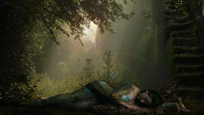 Mermaid-like woman with a teal tail lying on a blanket in a sunlit forest, a mossy stairway to the right.