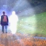 Person standing on a grassy path with two glowing translucent figures beside them in a misty outdoor scene.