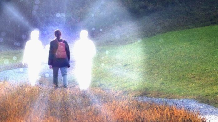 Person standing on a grassy path with two glowing translucent figures beside them in a misty outdoor scene.