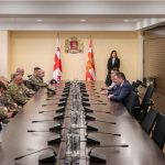 Long conference table with military personnel on the left and civilian officials on the right, two flags and a crest behind, plus a woman standing at the far end.
