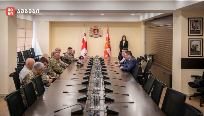 Long conference table with military personnel on the left and civilian officials on the right, two flags and a crest behind, plus a woman standing at the far end.